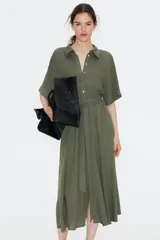 Vestido midi color verde oliva con cuello camisero, botonadura frontal y mangas raglán cortas. Tiene elástico fino en la parte posterior de la cintura, frunces adelante y atrás, y cinturón extraíble para atar.