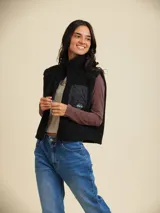 Chaleco negro de sherpa con cuello alto y cierre frontal con cremallera. Tiene un bolsillo vertical con cremallera en el pecho izquierdo y detalles en tejido liso en hombros y cuello.