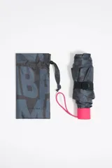 Paraguas plegable con funda cilíndrica de tela estampada con el logo de Bimba y Lola en tonos grises y negros. La base de la funda es de color fucsia y tiene un cordón de sujeción fucsia.