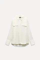 Camisa color blanco hueso de satén, con cuello solapa, manga larga, bolsillos con solapa en el delantero, bajo asimétrico y cierre frontal con botones.