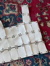 Top blanco de cuero genuino, confeccionado a mano con diseño de autor. La prenda está construida con tiras rectangulares de cuero unidas entre sí mediante ojales metálicos plateados, creando un efecto de cota de malla o armadura. Posee un tirante ancho en el hombro.