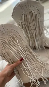 Botas texanas de caña media color crema, con punta fina, bordados y apliques de tachas. Presentan flecos largos en el lateral externo y cierre del lado interno.