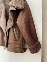 Campera marrón de gamuza sintética con cuello, puños y ruedo de piel sintética color marrón claro. Tiene cierre cruzado con cremallera y bolsillos laterales con cremallera.