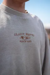 Buzo beige de corte holgado, tejido acanalado y cuello redondo. Estampado frontal con la frase "Glassy Waves Beach Life".