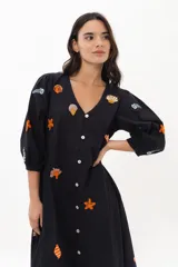 Vestido midi color arena con escote en V, mangas 3/4 abullonadas y cierre frontal con botones. Presenta bordados de motivos marinos.