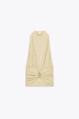 Top color crema con cuello halter, espalda descubierta, detalle de frunces y pieza metálica en forma de sol en el delantero.