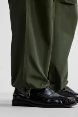 Pantalón verde militar de corte relaxed fit confeccionado en tejido técnico ligero. Cuenta con cintura elástica ajustable con cordón interior, bolsillos frontales y traseros con solapa, pliegues en las rodillas y bajo ajustable con elástico.