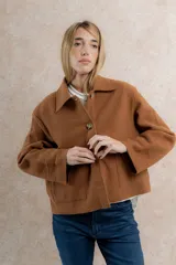 Chaqueta corta tipo blazer color marrón, confeccionada en tela de lana afieltrada. Tiene cuello camisero, silueta cuadrada, cierre frontal con botones y bolsillos delanteros.