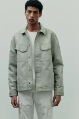 Sobrecamisa color arena de lona gruesa de algodón, corte recto, con cuello clásico, cierre frontal con botones a presión ocultos, canesú en la espalda, dos bolsillos superiores con solapa y botón a presión, bolsillos laterales discretos, mangas largas con puños abotonados y forro interno.