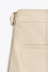 Falda pantalón color beige de tiro medio con diseño cruzado y cierre con botones. Cuenta con bolsillos falsos en la parte delantera y trasera, y cierre lateral con cremallera oculta.