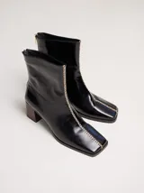 Botas de cuero charol negro con caña media y punta cuadrada. Cuentan con cierre trasero, tirador y bordado contrastante en el empeine. El taco es de madera y la suela de goma.