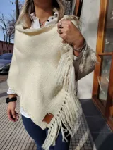 Chal color crema tejido a mano con lana fina y flecos. Presenta una etiqueta de cuero con el logo de la marca en una esquina.