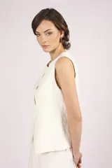 Chaleco negro sin mangas, con cuello redondo y cierre frontal de botones. Presenta detalles de encaje o apliques en los laterales y un corte ligeramente holgado.