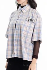 Camisa unisex de manga corta con estampado a cuadros en tonos verde, negro, blanco y morado. Presenta un corte boxy y un estampado en serigrafía en el pecho.