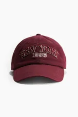 Gorra de béisbol color borgoña con bordado frontal de la frase "New York" y el número "1996" en blanco y rojo.