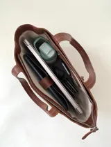 Cartera tipo tote de cuero graneado color negro, con textura uniforme y suave al tacto. Tiene manijas de 55 cm, bolsillo frontal con broche imantado, bolsillo trasero con cierre metálico, cierre principal metálico, forro interior y tres bolsillos internos (uno con cierre, uno plaqué y uno tipo sobre con solapa y botón).