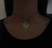 Gargantilla plateada con cadena de eslabones gruesos y cierre tipo toggle. Dije de corazón plateado con detalles ornamentales.
