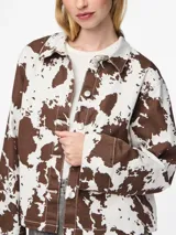 Chaqueta de jean con estampado animal print de vaca en tonos marrones y blancos, corte recto, cuello camisero y cierre frontal con botones.
