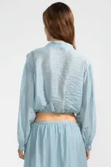 Blusa corta celeste en tejido arrugado liviano, con cuello mao, botones al frente y mangas abullonadas. Terminada con ruedo elastizado.