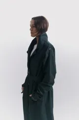 Trench largo color verde oscuro con doble botonadura, cuello clásico, solapas amplias, cinturón ajustable y charreteras en los hombros.