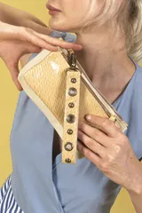 Sobre de mano color maíz con textura croco charol y detalles en cuero graneado color offwhite. Tiene doble cierre dorado, correa muñequera desmontable con tachas y forro textil liso.