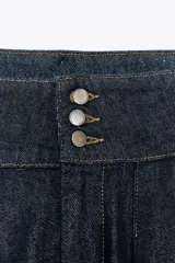 Pantalón bombacho de denim azul oscuro, tiro alto, con bolsillos delanteros y puños con botón en los tobillos. Cierre frontal con cremallera y botones metálicos.