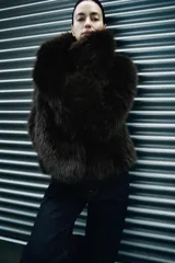 Abrigo corto marrón de pelo sintético, con cuello solapa, manga larga, bolsillos delanteros, forro interior y cierre frontal con botón.