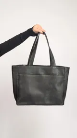 Cartera tipo tote de cuero graneado color negro, con textura uniforme y suave al tacto. Tiene manijas de 55 cm, bolsillo frontal con broche imantado, bolsillo trasero con cierre metálico, cierre principal metálico, forro interior y tres bolsillos internos (uno con cierre, uno plaqué y uno tipo sobre con solapa y botón).