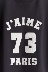 Buzo negro de corte oversized con interior cepillado suave, cuello redondo y mangas largas. Presenta un estampado frontal con el texto "J'AIME 73 PARIS" en color blanco.