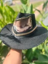 Sombrero de ala ancha tejido en hilo de coco y sintético, color marrón claro. La copa está cubierta con encaje naranja quemado con patrón floral. Tiene una cinta de cuero marrón oscuro y un aplique decorativo de hoja de gamuza con incrustaciones de pedrería.