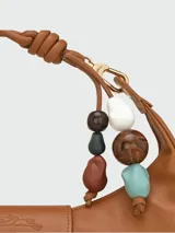 Bolso hobo de cuero color marrón, con cierre de solapa con botón a presión y correa corta para llevar al hombro. Incluye charm colgante con cuentas de madera y piedra.