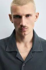 Camisa de denim de algodón, color beige claro, con corte relajado, cuello bowling y manga corta. Presenta cierre frontal con botones a presión y un pequeño bordado a tono en el pecho.