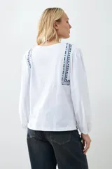 Remera blanca de estilo boho con bordados en el escote y mangas largas. Presenta escote en V con lazos y borlas. Corte recto y holgado.
