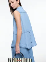 Camisa sin mangas en denim color celeste, confeccionada en 100% lyocell. De corte recto y largo medio, presenta cartera escondida en el frente y un bolsillo plaqué en el pecho. En la espalda, tiene botones decorativos alineados en abertura vertical y pespuntes a contratono.