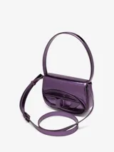 Bolso bandolera Diesel modelo 1DR, color violeta metalizado, con asa corta rígida y correa larga extraíble. Presenta solapa con cierre magnético y logo ovalado "D" en relieve.