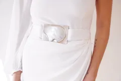 Vestido largo blanco de seda, con un hombro descubierto, manga larga abullonada y falda irregular con drapeado lateral. Incluye cinturón blanco con hebilla de alpaca y nácar.
