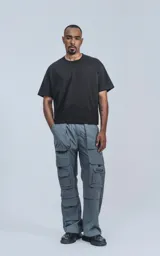 Pantalón cargo técnico gris oscuro, de silueta oversize y tejido liviano. Presenta múltiples bolsillos cargo con solapa en las piernas, uno de los cuales tiene una pequeña etiqueta negra con texto.