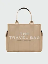 Bolso tote beige de lona con asas de mano negras y estampado frontal con la leyenda "THE TRAVEL BAG MARC JACOBS". Cuenta con bolsillos laterales externos.