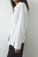 Camisa blanca de cuello solapa y manga larga. Detalle de pliegues con botones. Cierre frontal con botones.
