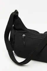 Cartera tipo shoulder color beige, de diseño utilitario y formato curvo. Confeccionada en material liviano y resistente, cuenta con correa regulable para llevar al hombro o cruzada. Presenta múltiples bolsillos con cierre en el frente y laterales, y un parche con el logo "Piece & Cake" en el frente.