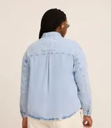 Camisa de jean de algodón, color celeste claro lavado, con cuello tradicional, manga larga y bolsillo de parche en el pecho. Presenta un cierre frontal con botones metálicos.