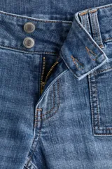 Jeans de denim azul medio con ligero efecto desgastado y roturas a la altura de las rodillas. Presenta un corte ajustado en muslos y acampanado desde la rodilla. El tiro es bajo, con pretina prolongada que se cierra con dos botones visibles. Incluye bolsillos delanteros tipo parche y bolsillos traseros.