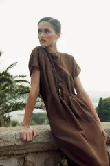 Vestido midi color marrón confeccionado en hilatura con mezcla de algodón y lino. Tiene cuello redondo, manga corta caída y detalle de pliegues en la cintura. Cierre en la espalda con abertura y botón.