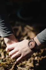 Reloj de pulsera con caja, esfera y malla de madera de sándalo rojo, con marcadores y manecillas negras. Presenta un diseño circular con corona lateral.