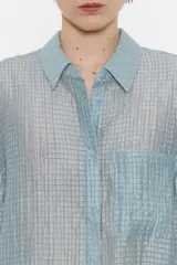 Camisa larga de viscosa con textura, de corte recto, con cuello camisero, manga larga de puño con botón y cierre frontal con abotonadura. Presenta bolsillo de plastrón en el pecho y nudo decorativo en la parte trasera para dar volumen.