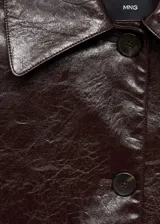 Chaqueta corta color marrón oscuro de poliuretano con textura, cuello camisero, manga larga con puños abotonados, dos bolsillos de solapa en la parte delantera y cierre delantero con botones.