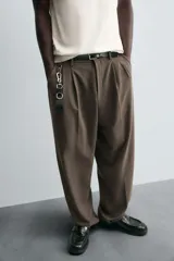 Pantalón negro de corte ancho con cintura elástica, pliegues frontales, bolsillos laterales y traseros de vivo. Tiene detalle de hebilla metálica en la trabilla y cierre frontal con cremallera y botón.