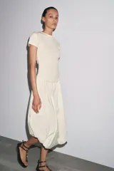 Vestido largo color crema, con cuello redondo y mangas cortas. Presenta corte a la altura de la cintura y falda amplia con terminación redondeada tipo globo.