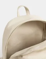 Mochila de cuerina color crudo con bolsillo frontal con cierre y charm decorativo con cierre. Cuenta con correas regulables y manija superior.