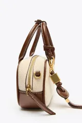 Bolso mini color crudo con detalles en marrón. Tiene asa de mano y doble asa bandolera extraíble, una en tejido tipo canvas y otra con pespunte. Incluye charm con piezas metálicas y bolsillo interior con cremallera.